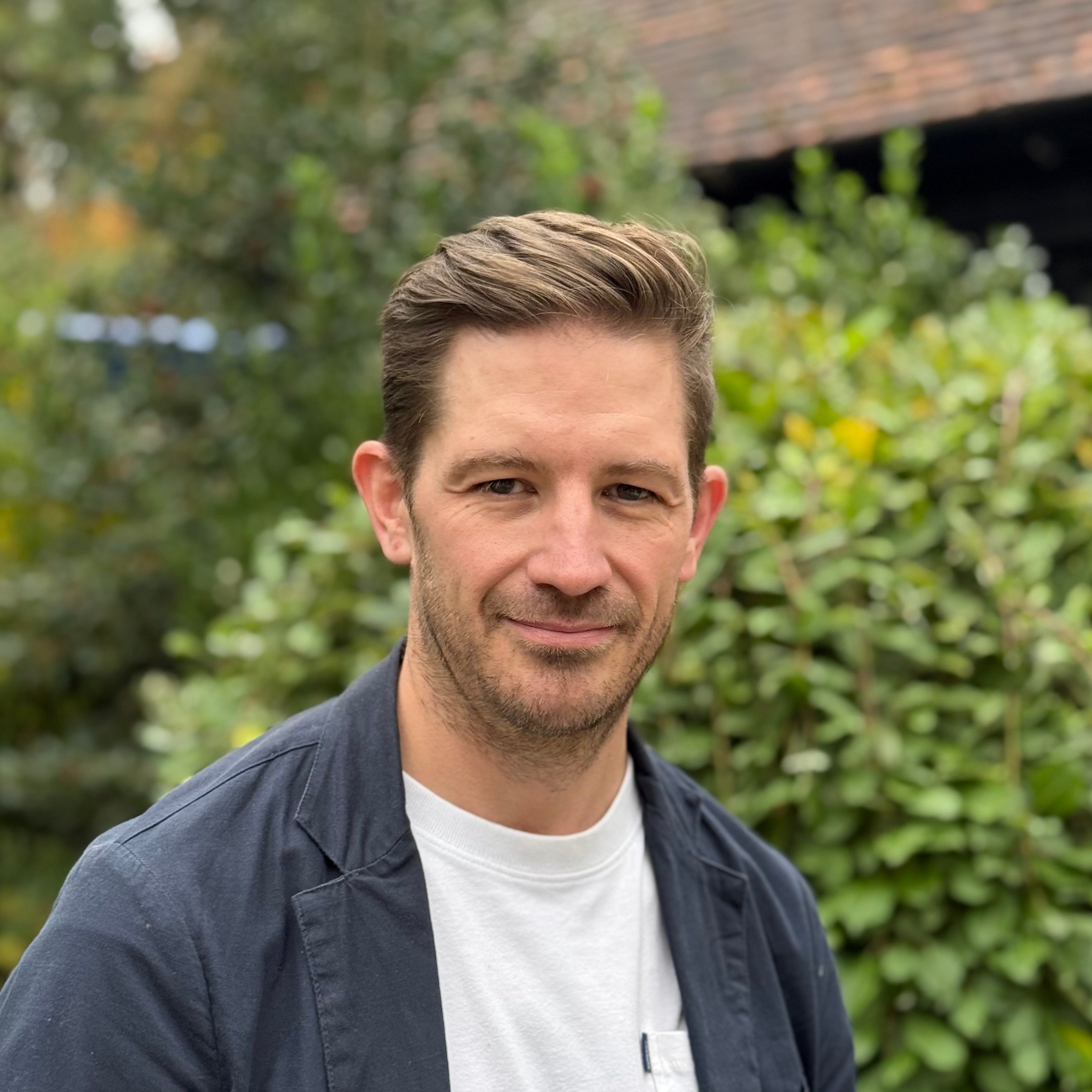 Image of Mark Kelly wearing white t-shirt and navy blazer with a greenery backdrop. Image of Mark Kelly wearing white t-shirt and navy blazer with a greenery backdrop.